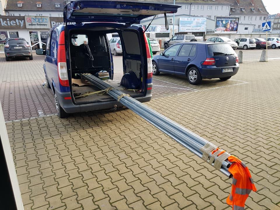 Transport rur instalacyjnych zabezpieczonych pomarańczową kamizelką odblaskową wystających z otwartego bagażnika niebieskiego busa na parkingu.