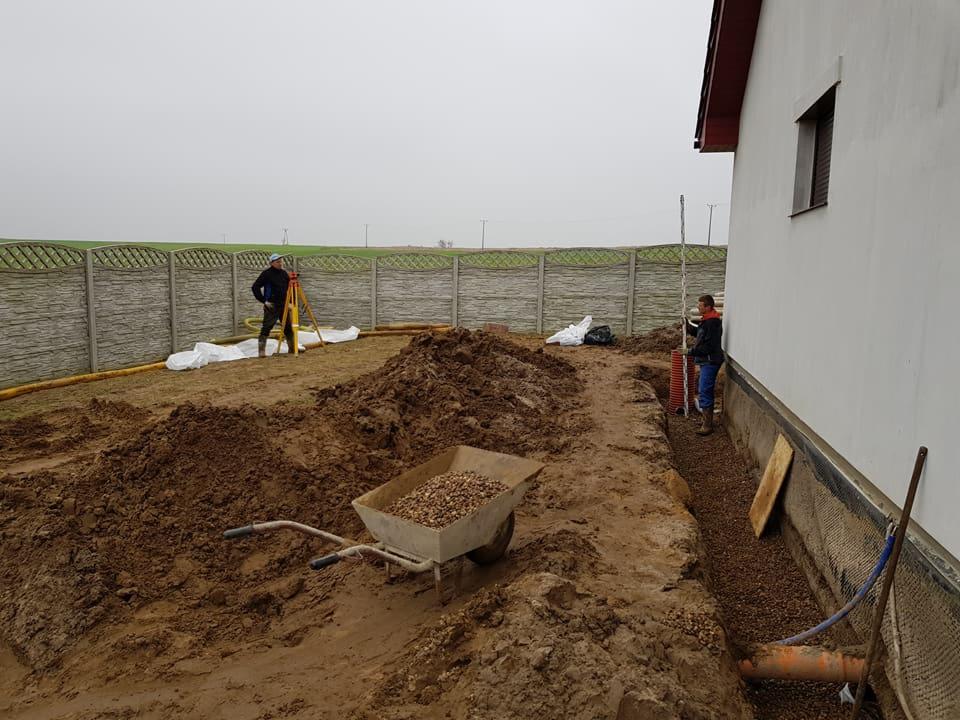 Działka w trakcie prac ziemnych pod budowę, widoczne wykopy, gruz w taczce, pracownicy z niwelatorem i rurami kanalizacyjnymi w wykopie przy budynku.