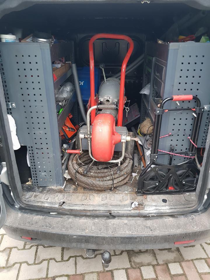 Wnętrze samochodu serwisowego z widoczną maszyną do czyszczenia kanalizacji z czerwoną obudową i spiralnym wężem, obok regały z narzędziami i akcesoriami hydraulicznymi.