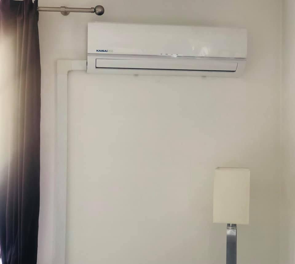 Biały klimatyzator marki Kaisai zamontowany na ścianie w jasnym pomieszczeniu, z widoczną instalacją rur i fragmentem zasłony oraz lampy.