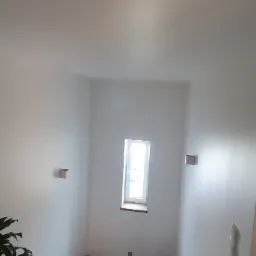 Schody drewniane prowadzące do pomieszczenia z małym oknem, białe ściany, roślina doniczkowa w lewym dolnym rogu, oświetlenie punktowe na suficie i kinkiet na ścianie.