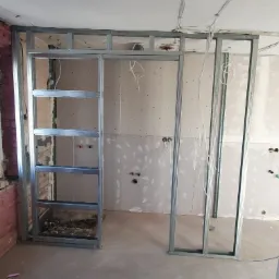 Wnętrze w trakcie remontu z widoczną metalową konstrukcją ścianki działowej, częściowo obłożoną płytami, odsłonięte przewody elektryczne i surowy sufit z widocznymi śladami starych instalacji.