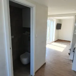 Widok na wykończoną łazienkę z toaletą i drewnopodobną podłogą, widok z korytarza z panelami podłogowymi, jasne ściany, białe drzwi przesuwne.