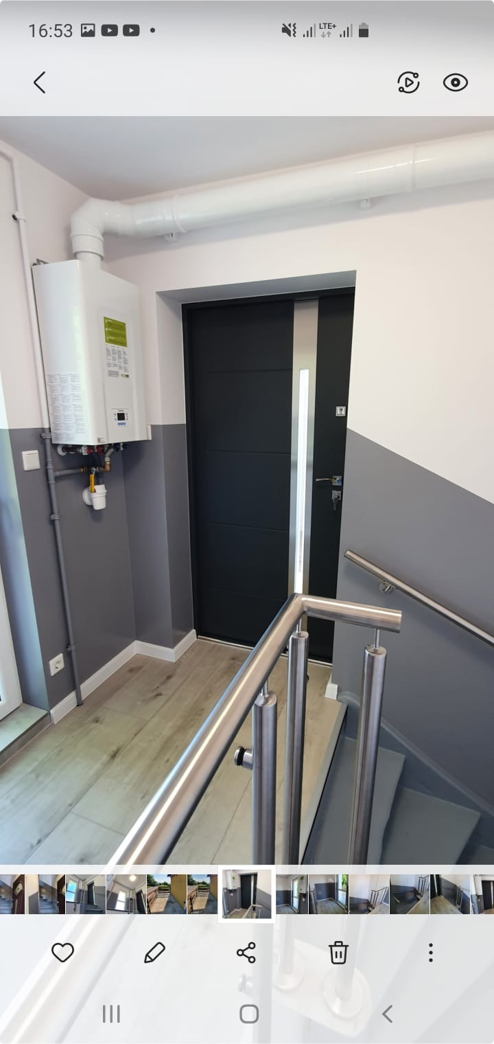 Wnętrze przedpokoju z szaro-białymi ścianami, drewnianą podłogą, grzejnikiem gazowym i nowoczesnymi, czarnymi drzwiami z metalowym akcentem oraz metalową barierką schodową.