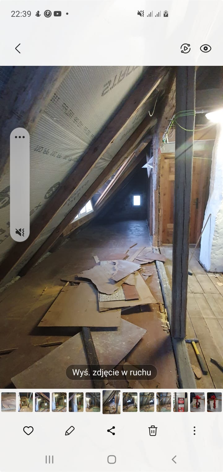 Poddasze w trakcie remontu, widoczne drewniane belki stropowe, fragmenty płyt na podłodze, okno dachowe w oddali i narzędzia pracy.