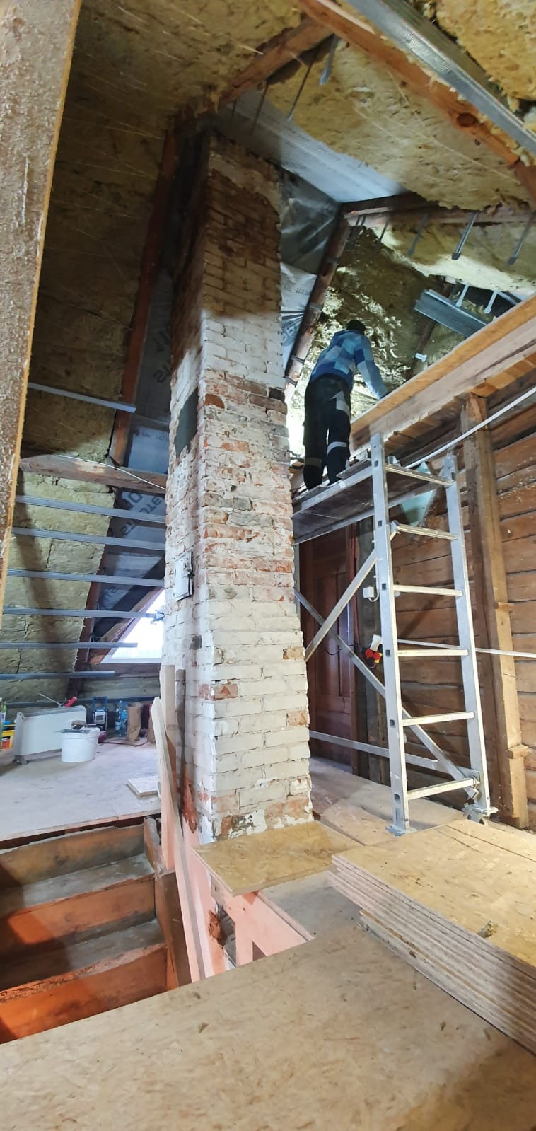 Poddasze w trakcie remontu: ocieplenie, stara ceglana kolumna kominowa, pracownik na rusztowaniu, widoczne materiały budowlane i przygotowane płyty OSB.
