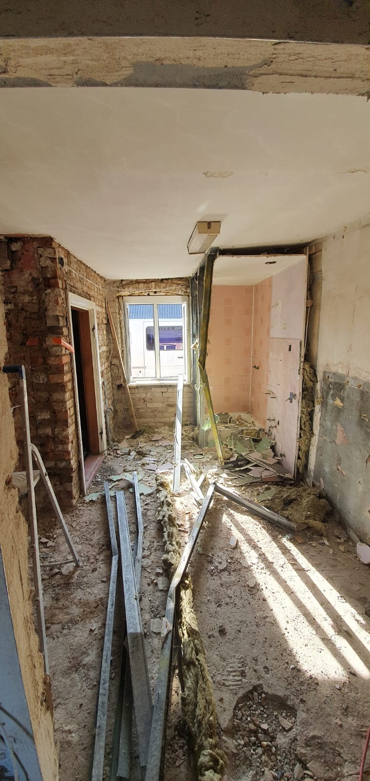 Wnętrze w trakcie remontu: odsłonięta cegła, resztki płytek w łazience, gruz na podłodze, widoczne profile metalowe i drabina.