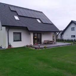 Nowoczesny dom jednorodzinny z ciemną dachówką i panelami słonecznymi na dachu, widok z zadbanego trawnika, w tle kolejne domy.