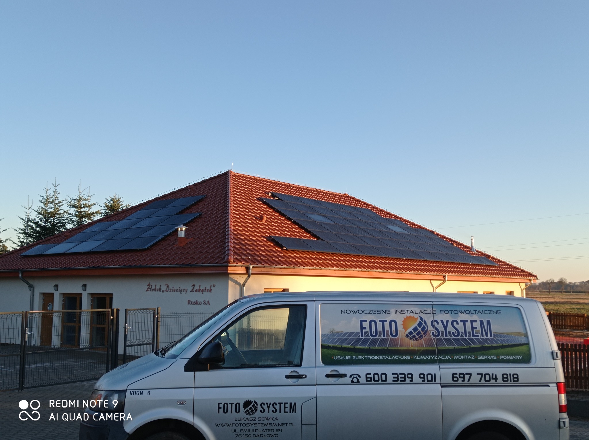 Budynek Żłobka Dziecięcego Zakątka z panelami słonecznymi na dachu, częściowo zasłonięty przez srebrny samochód dostawczy firmy Foto System z widocznym logo i numerami telefonów.