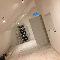 Poddasze w trakcie przygotowania do wykończenia: świeżo zagruntowane ściany, rozstawiony ruszt, widoczne ślady szpachlowania i resztki materiałów na podłodze, okrągłe okno w szczycie ściany.