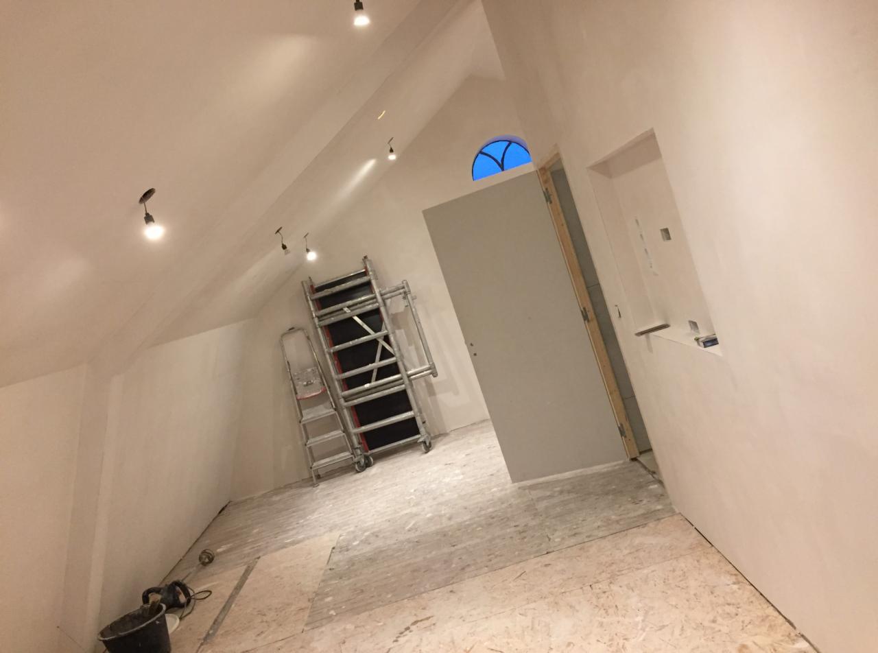 Poddasze w trakcie przygotowania do wykończenia: świeżo zagruntowane ściany, rozstawiony ruszt, widoczne ślady szpachlowania i resztki materiałów na podłodze, okrągłe okno w szczycie ściany.