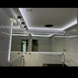 Nowoczesna łazienka w trakcie wykańczania, z białymi marmurkowymi płytkami, podświetlanym sufitem z punktowymi lampami i dużym lustrem odbijającym wnętrze.