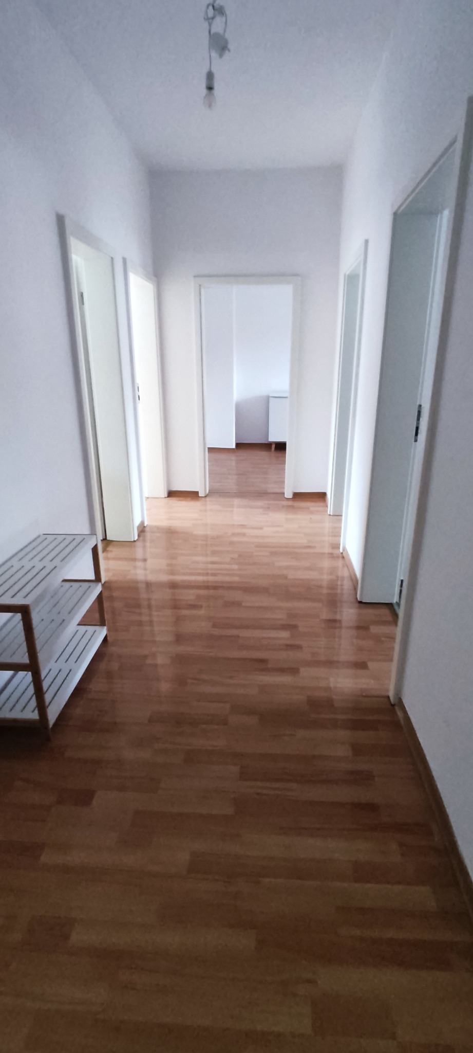 Korytarz z drewnianą podłogą w jasnym mieszkaniu, widoczne trzy pary białych drzwi, białe ściany i biały regał na buty.