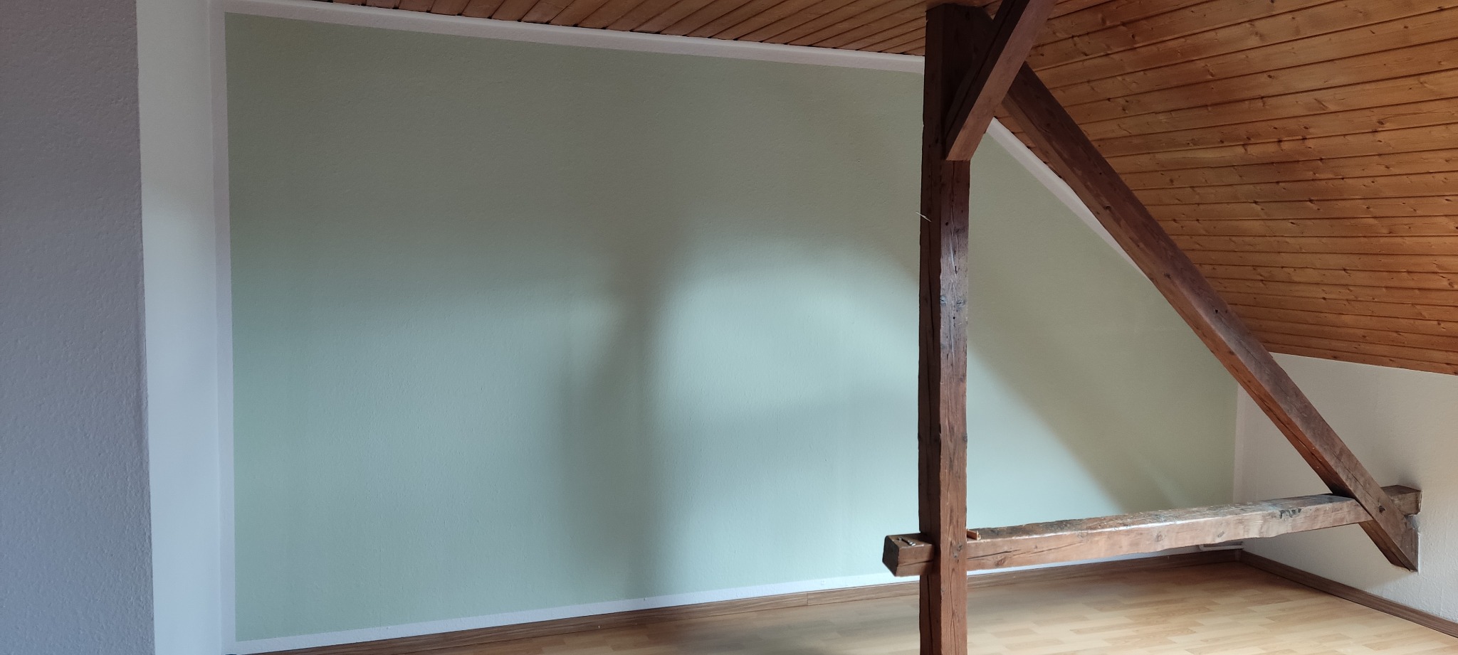 Pomalowana na jasnozielono ściana pod skosem dachu z drewnianą konstrukcją nośną, widok na fragment poddasza z drewnianą boazerią.