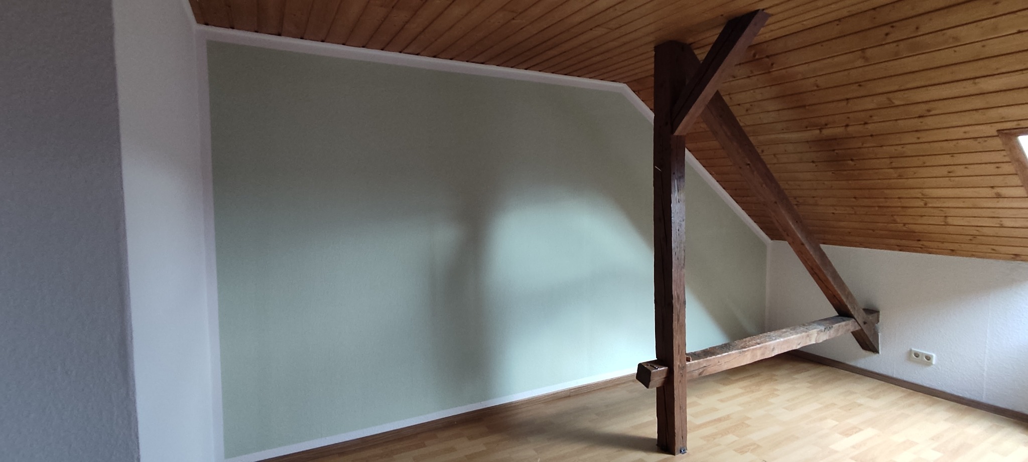 Pomalowane na jasnozielono ściany poddasza z drewnianą więźbą dachową, widoczny fragment drewnianego sufitu i laminowanej podłogi.