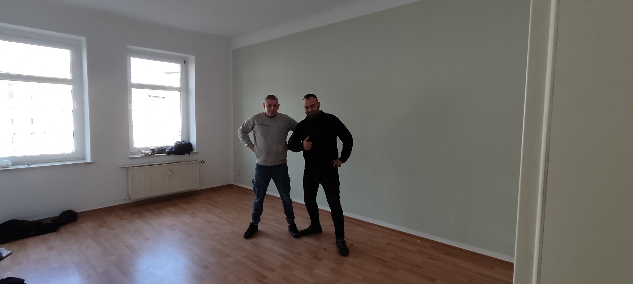 Dwaj mężczyźni pozują w pustym, świeżo pomalowanym pokoju z jasnymi, gładkimi ścianami w odcieniu seledynu i drewnianą podłogą; jeden z nich pokazuje kciuk w górę.