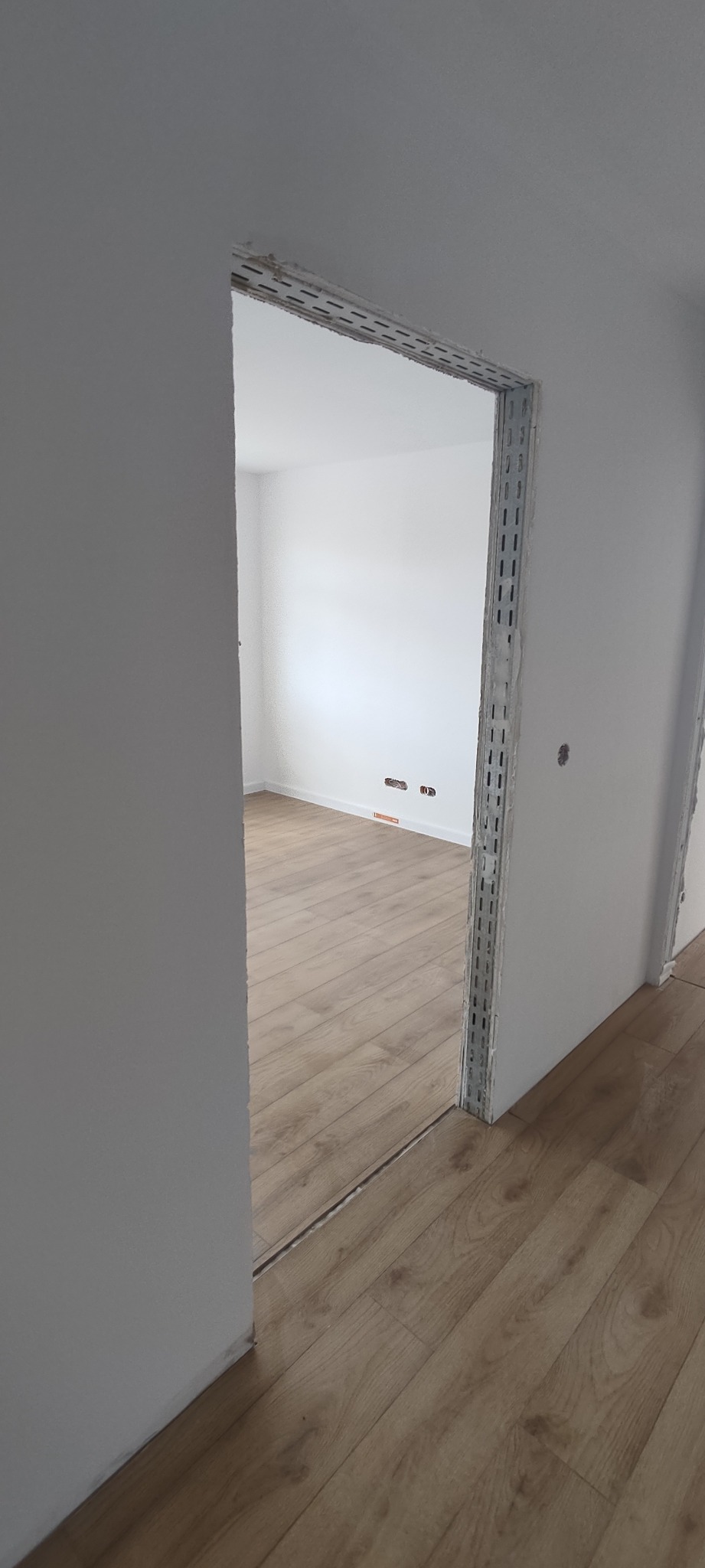 Przygotowanie do montażu ościeżnicy drzwiowej w surowym stanie, widoczne profile montażowe i surowa ściana, parkiet w odcieniach brązu.