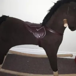 Ciemnobrązowy koń na biegunach z brązowym siodłem, widoczny z boku, stoi na dywanie. W tle biała ściana i czerwony pojemnik na narzędzia.