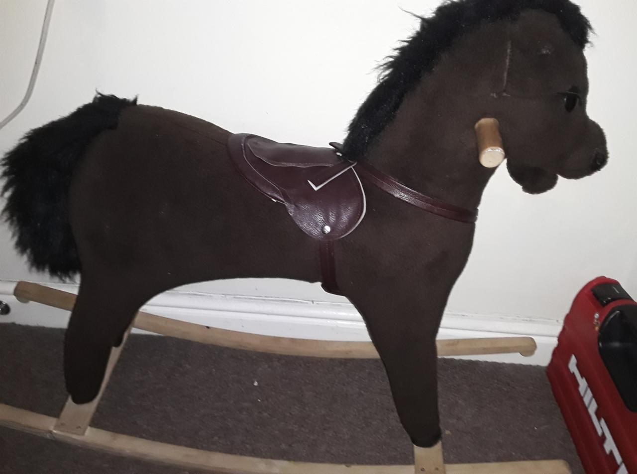 Ciemnobrązowy koń na biegunach z brązowym siodłem, widoczny z boku, stoi na dywanie. W tle biała ściana i czerwony pojemnik na narzędzia.