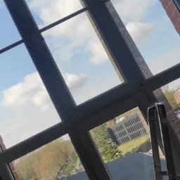 Widok z okna z czystymi szybami, przez które widać błękitne niebo z chmurami i budynki na zewnątrz; drabina oparta o ramę okienną.