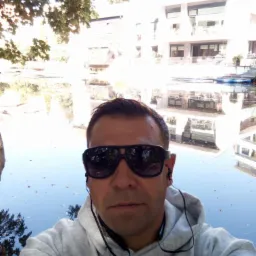 Mężczyzna w okularach przeciwsłonecznych robi selfie na tle jeziora i budynku z balkonami w słoneczny dzień. W tle widoczne drzewa i odbicia w wodzie.