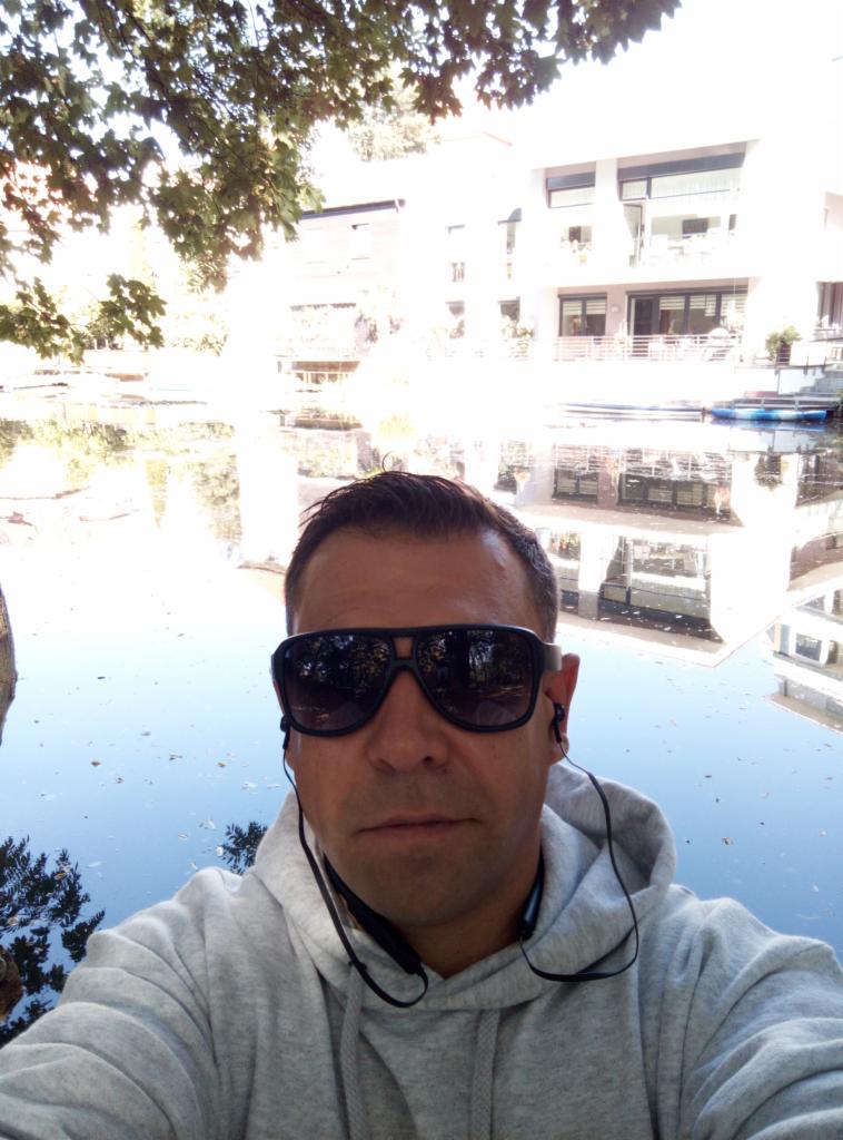 Mężczyzna w okularach przeciwsłonecznych robi selfie na tle jeziora i budynku z balkonami w słoneczny dzień. W tle widoczne drzewa i odbicia w wodzie.