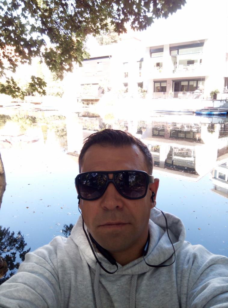 Mężczyzna w okularach przeciwsłonecznych, szarej bluzie z kapturem i słuchawkami na tle jeziora i budynku odbijającego się w wodzie.