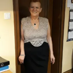 Uśmiechnięta kobieta w koronkowej bluzce i czarnej spódnicy, stojąca w drzwiach, z kalendarzem i biurkiem w tle. Detal: bransoletka na lewym nadgarstku.