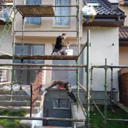 Dwaj pracownicy na rusztowaniu podczas prac elewacyjnych. Jeden nakłada zaprawę, drugi pracuje przy wejściu do piwnicy. Okna zabezpieczone folią malarską.