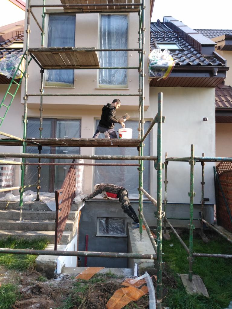Dwaj pracownicy na rusztowaniu podczas prac elewacyjnych. Jeden nakłada zaprawę, drugi pracuje przy wejściu do piwnicy. Okna zabezpieczone folią malarską.