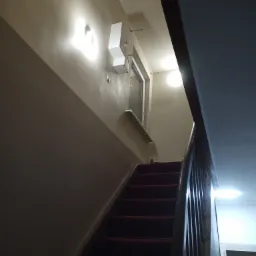 Wnętrze klatki schodowej z czerwonym dywanem na schodach, ściany pomalowane w dwóch odcieniach beżu, oświetlenie punktowe na ścianie i suficie.