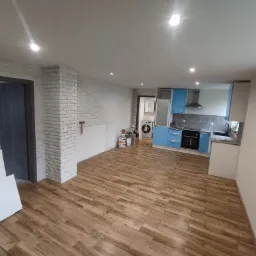 Wnętrze mieszkania w trakcie remontu: położona podłoga z paneli imitujących drewno, ściana z białej cegły, niebieska zabudowa kuchenna, widoczne puszki z farbą i elementy wyposażenia.