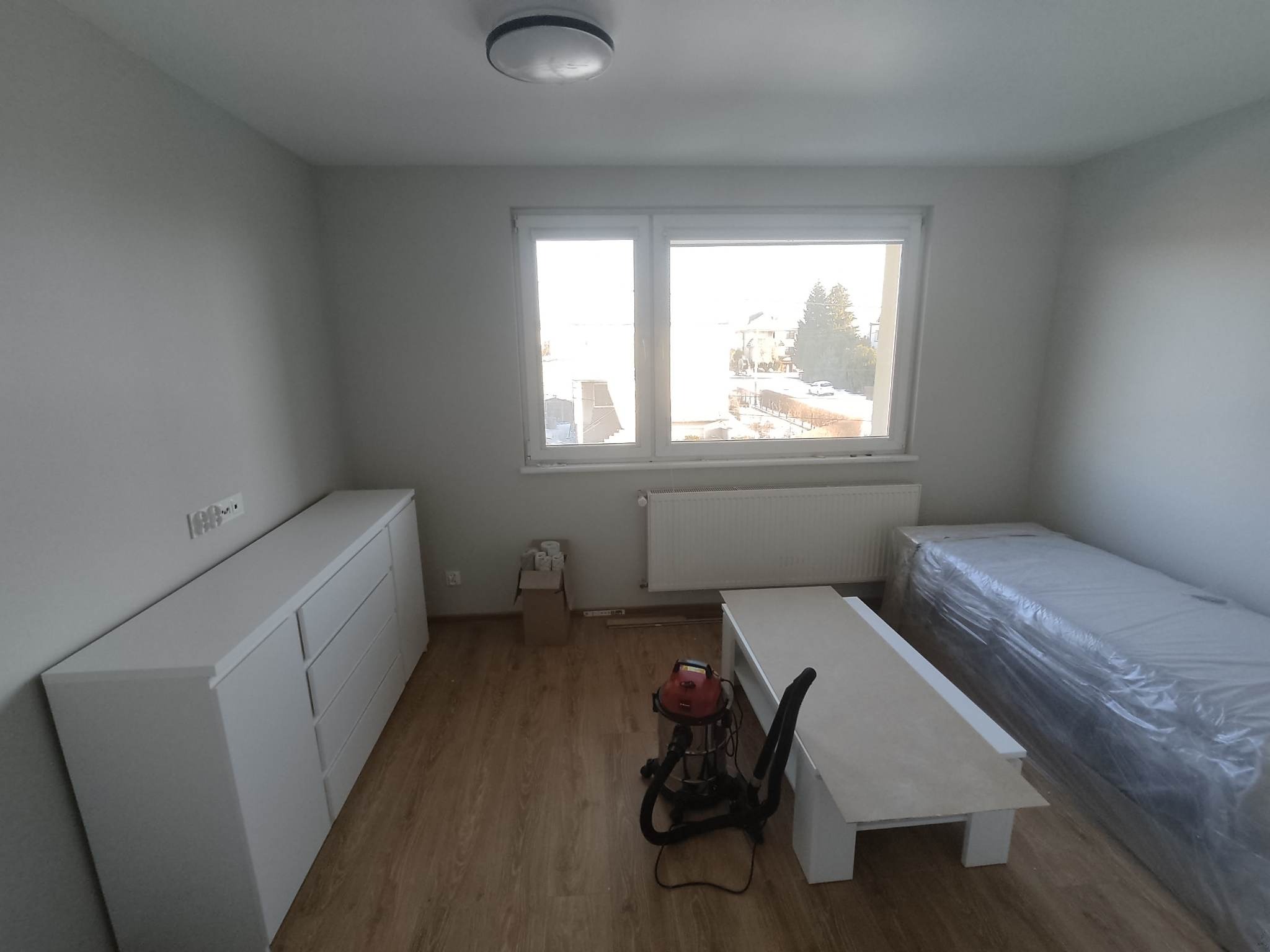 Wnętrze pokoju po remoncie: białe meble, okno z widokiem na zewnątrz, odkurzacz przemysłowy i łóżko zabezpieczone folią ochronną.