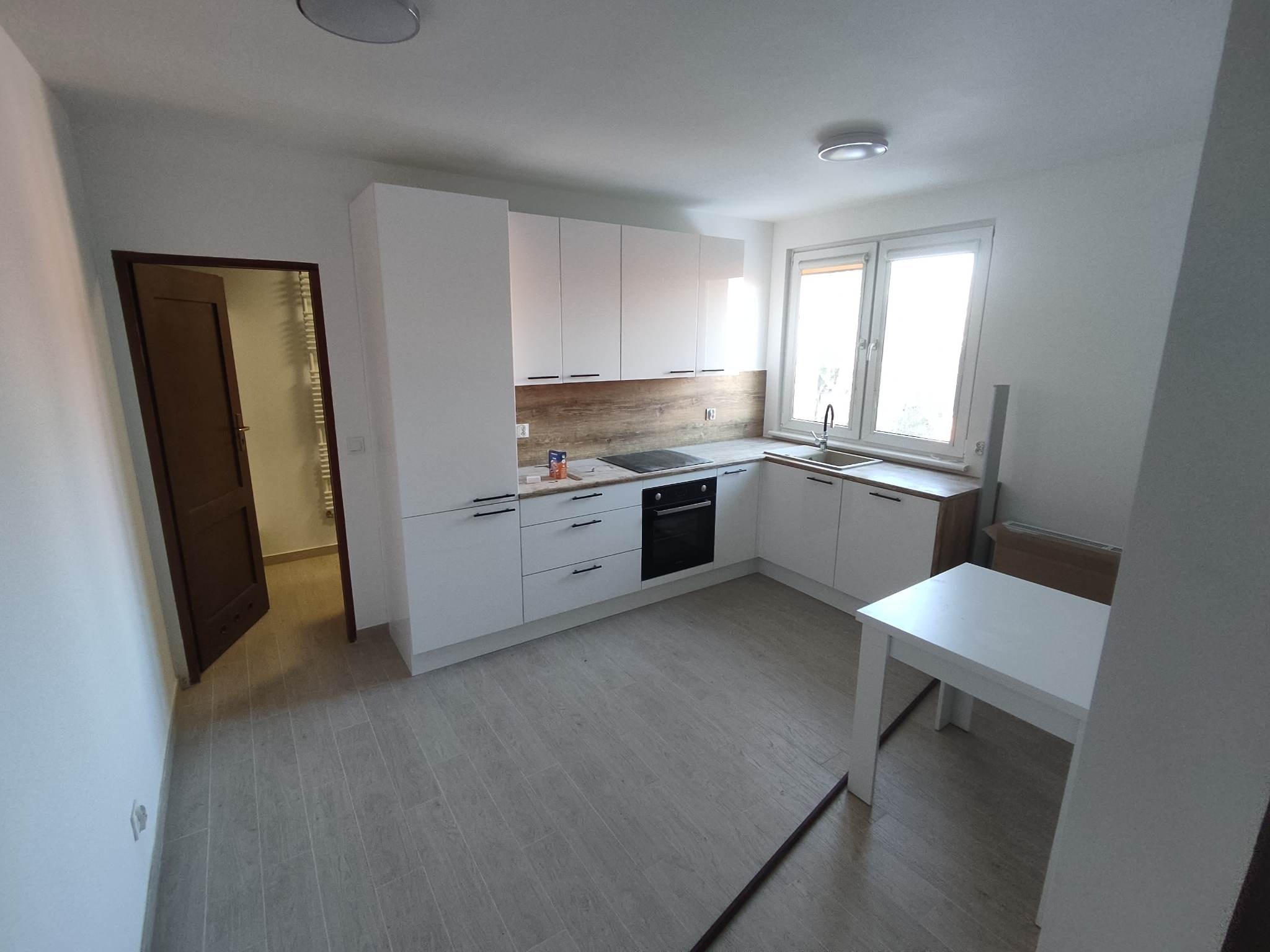 Jasna kuchnia z białymi szafkami, drewnopodobnym panelem nad blatem i szarymi płytkami podłogowymi. Widoczny stół i otwarte drzwi do innego pomieszczenia.