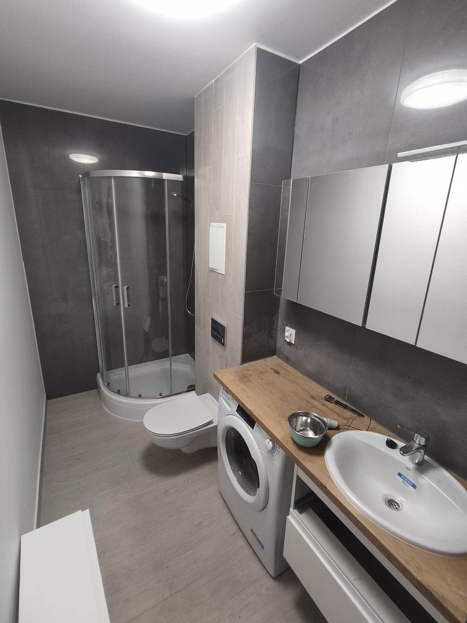 Nowoczesna łazienka w szarościach z drewnianym blatem, kabina prysznicowa z zaokrąglonymi drzwiami, umywalka z armaturą Roca Rodeo i metalowa miska obok pralki.