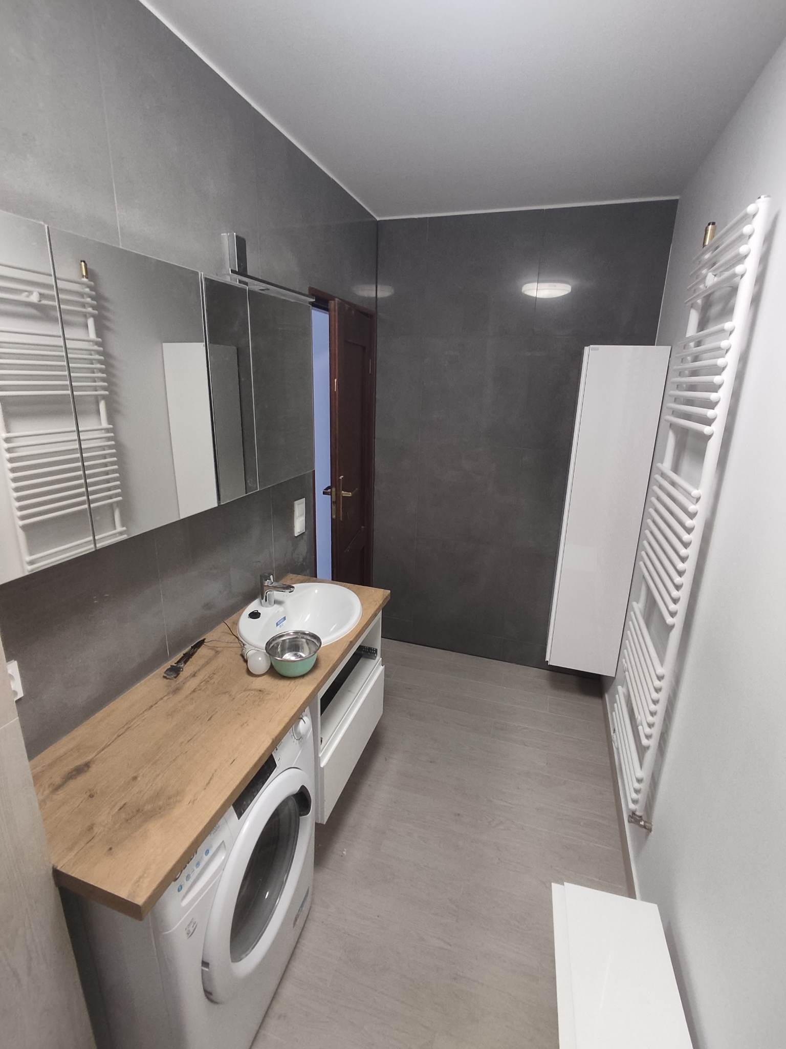 Szara łazienka z umywalką na blacie z drewnopodobnego laminatu, pralką pod blatem, grzejnikiem drabinkowym i lustrem w zabudowie. Na blacie leżą narzędzia i miska.