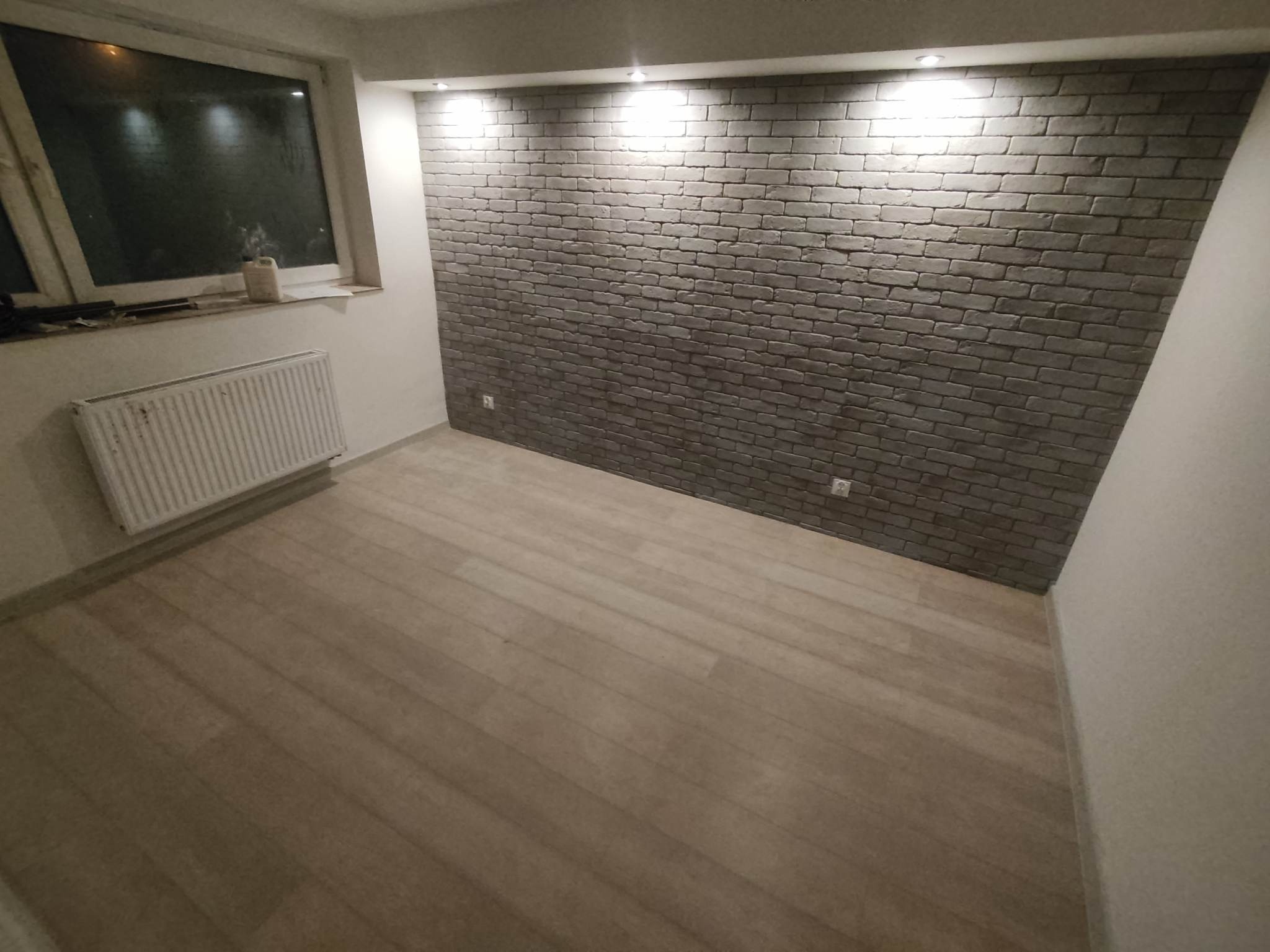 Ściana z szarej cegły dekoracyjnej w nowym pokoju z panelami podłogowymi i grzejnikiem pod oknem.