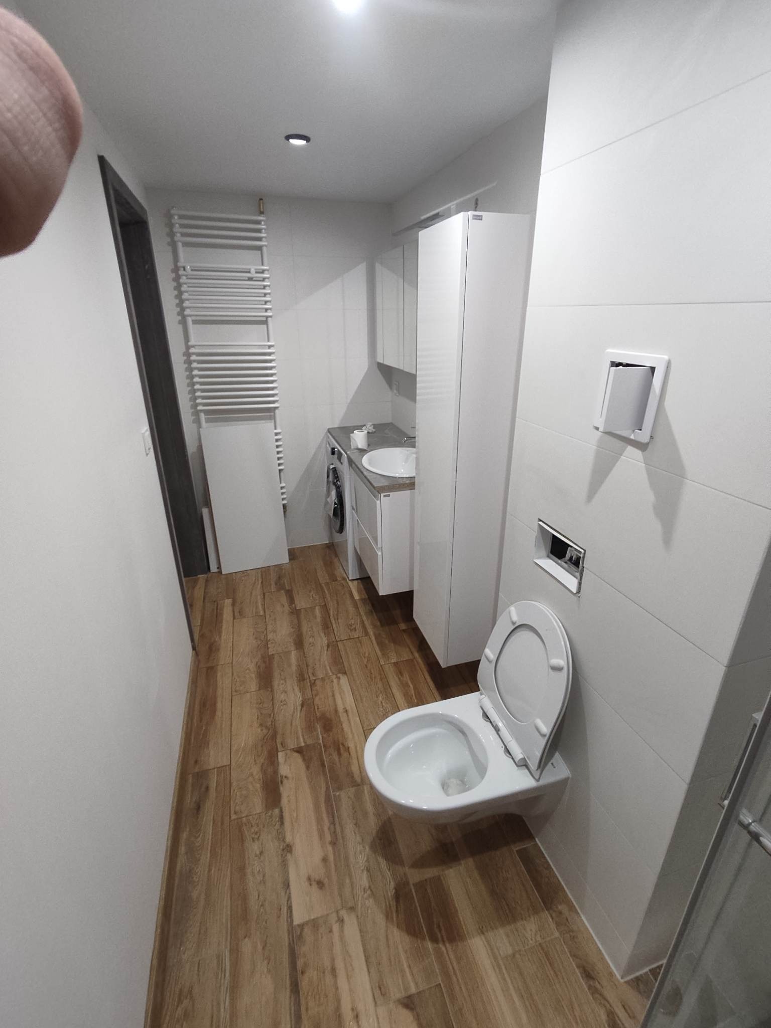 Nowoczesna łazienka w bieli i brązie z drewnopodobnymi płytkami podłogowymi, wiszącą toaletą z otwartą klapą, grzejnikiem drabinkowym i szafką z umywalką.