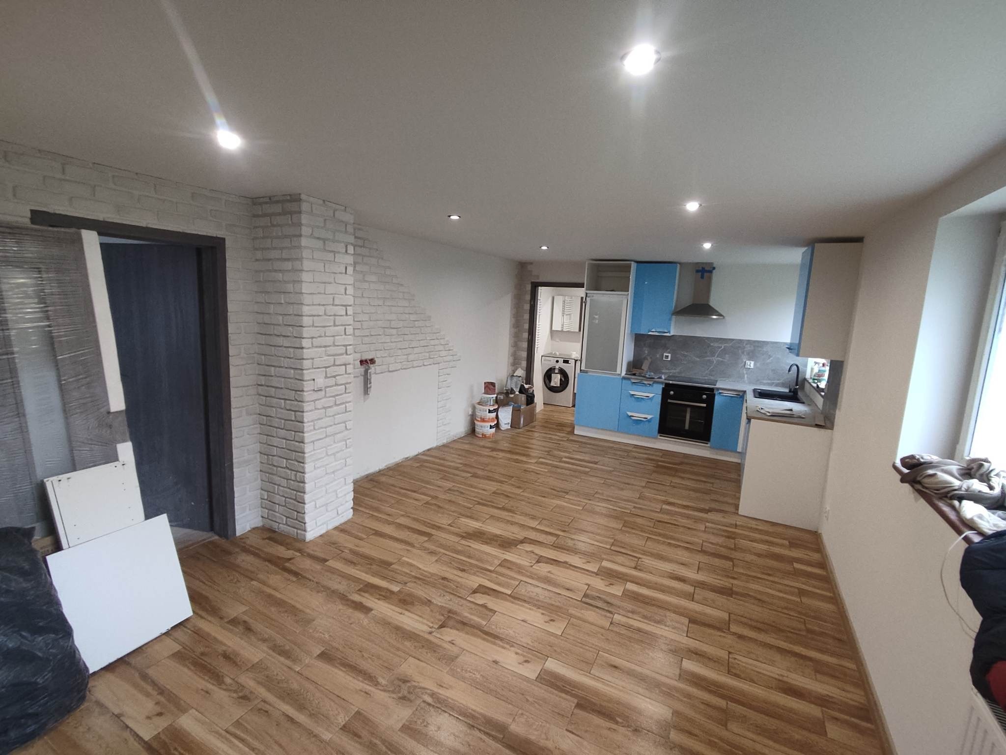 Wnętrze mieszkania w trakcie remontu: położona podłoga z paneli imitujących drewno, ściana z białej cegły, niebieska zabudowa kuchenna, widoczne puszki z farbą i elementy wyposażenia.