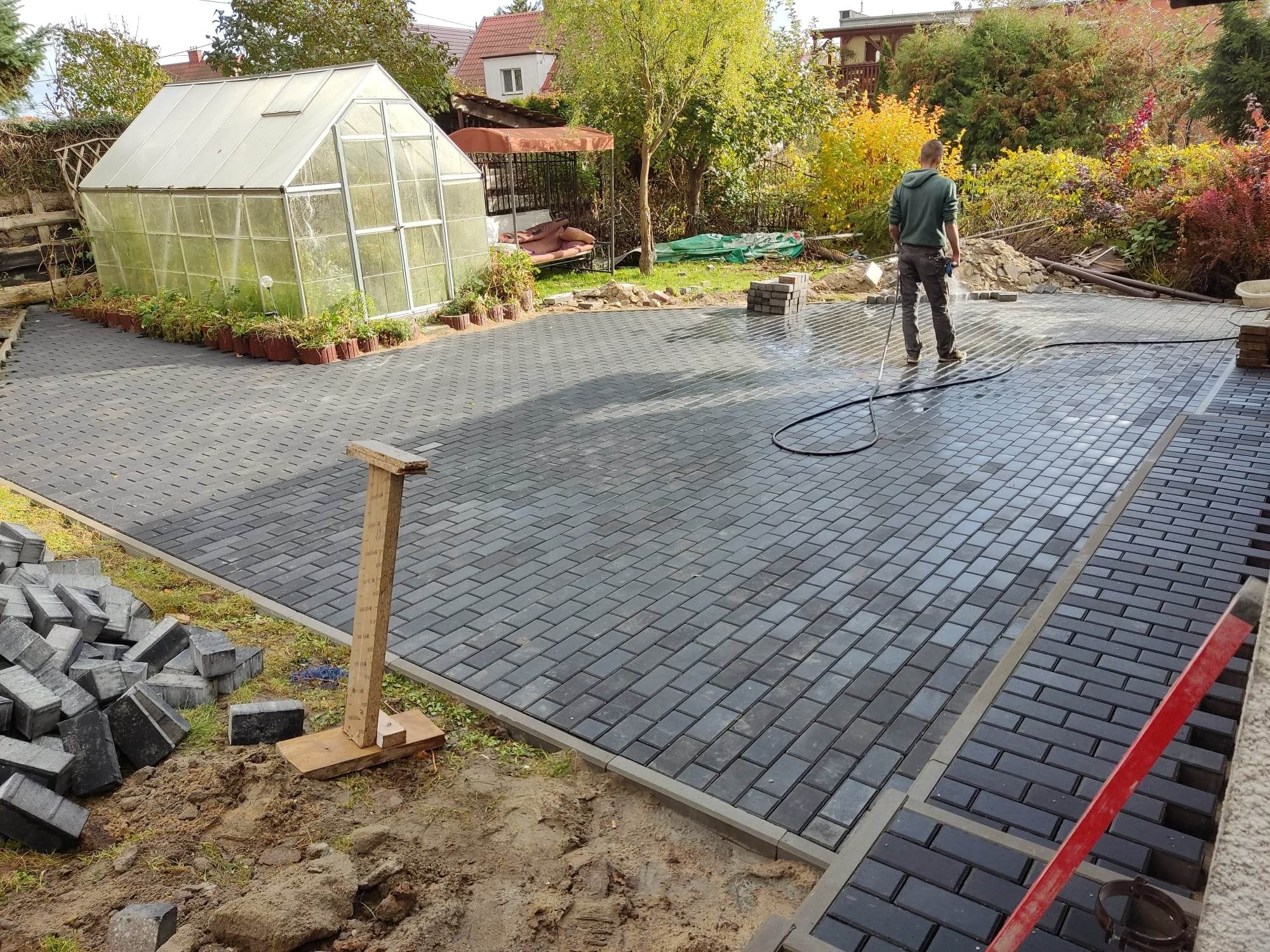 Mężczyzna myje wodą nowo ułożoną, szarą kostkę brukową na posesji z widoczną szklarnią i zielenią w tle, część kostki jeszcze nieułożona.