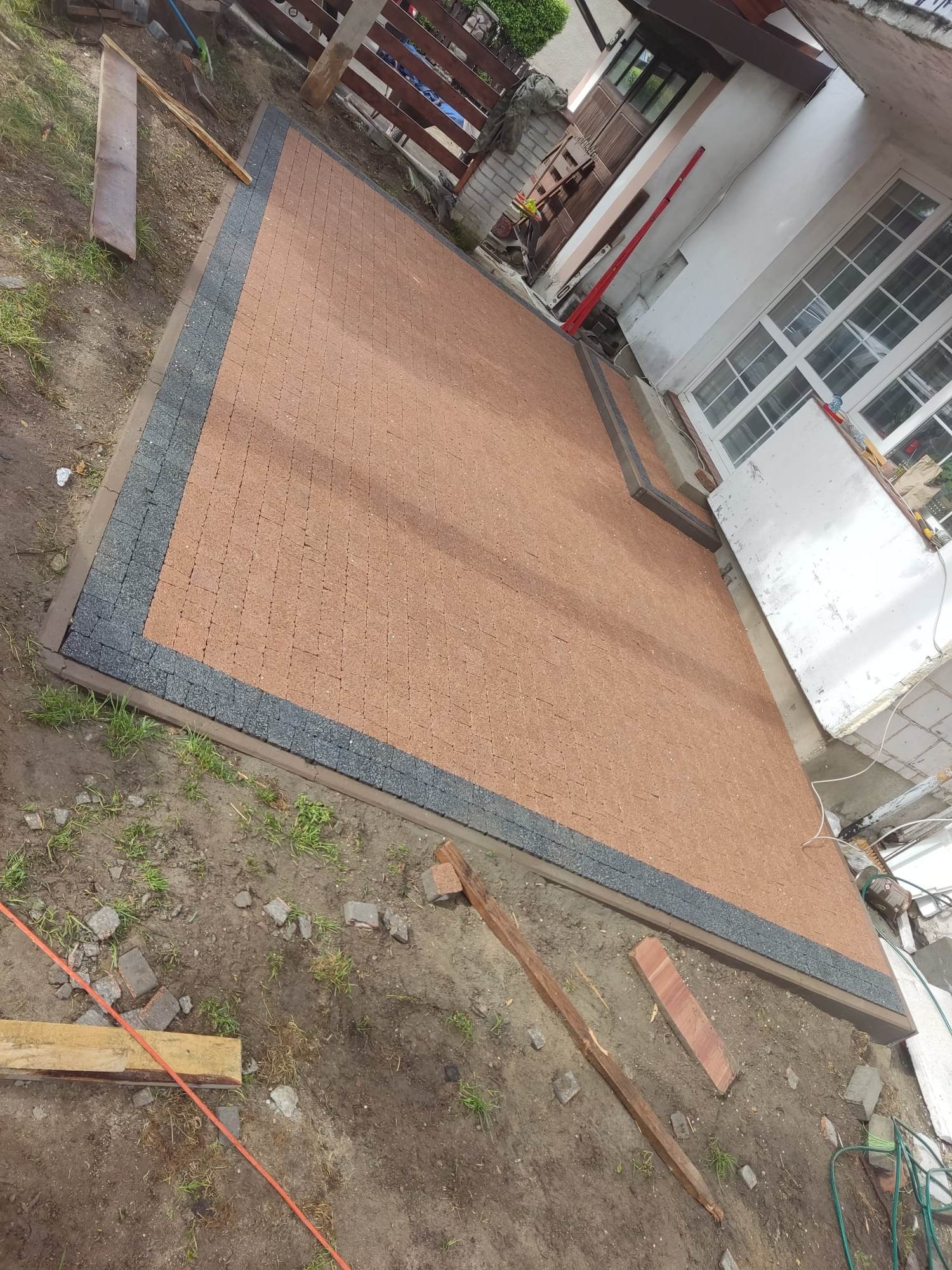 Nowo ułożona, brązowa kostka brukowa z ciemnoszarym obramowaniem, przylegająca do ściany budynku z widocznymi oknami i elementami drewnianymi, otoczona ziemią i resztkami materiałów budowlanych.