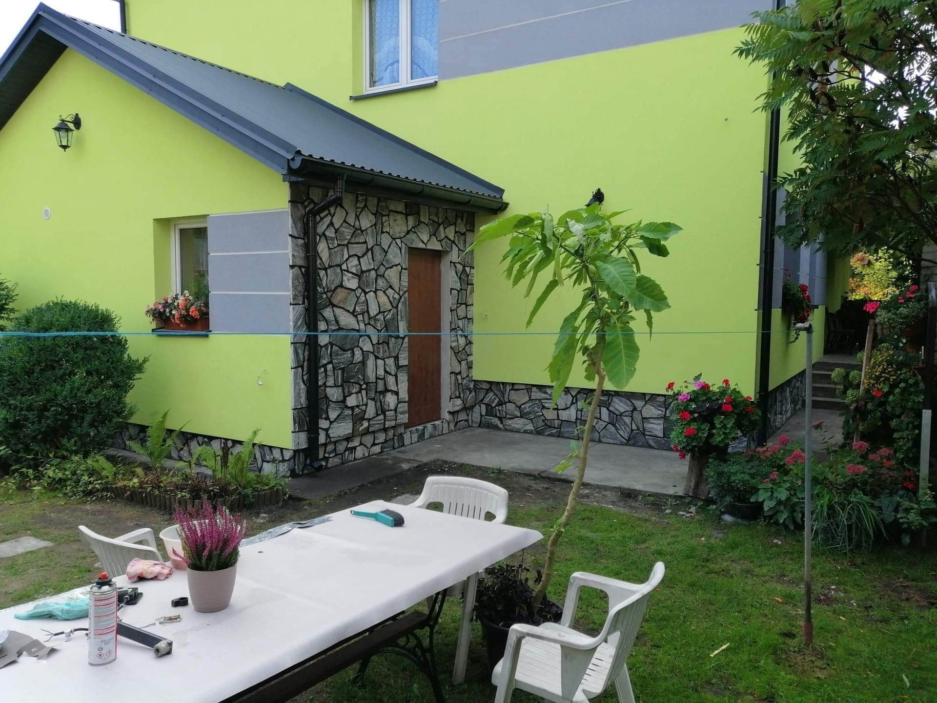 Dom z elewacją w odcieniach zieleni i kamienną okładziną przy wejściu, widoczny stół z narzędziami i kwiatami w ogrodzie.