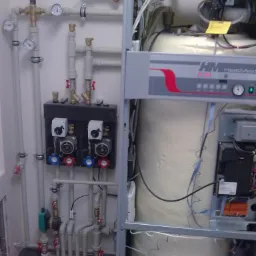 Kocioł grzewczy z otwartą obudową, widoczne elementy instalacji hydraulicznej, rury, zawory, manometry oraz pompy obiegowe na ścianie.