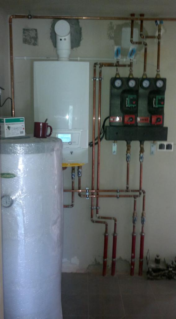 Biały piec grzewczy z miedzianymi rurami, zbiornik wody owinięty folią, trzy czarne panele sterujące z manometrami, czerwony kubek na zbiorniku, instalacja w surowym pomieszczeniu.