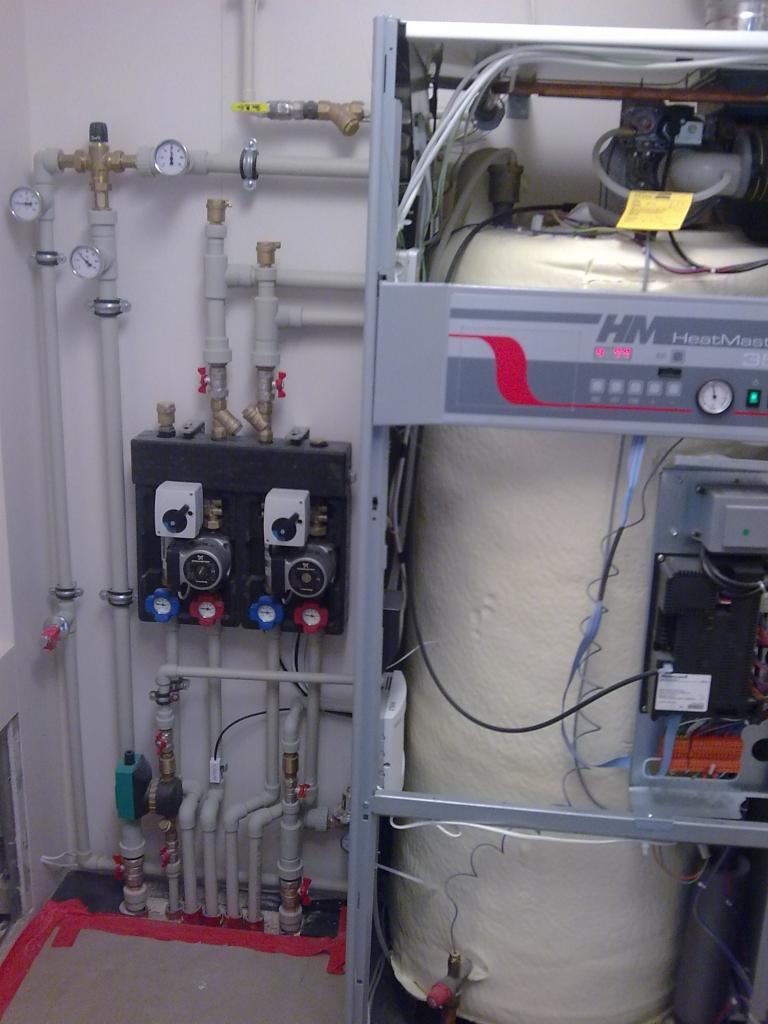 Kocioł grzewczy z otwartą obudową, widoczne elementy instalacji hydraulicznej, rury, zawory, manometry oraz pompy obiegowe na ścianie.