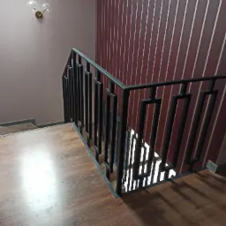 Nowoczesna, czarna metalowa balustrada schodowa o geometrycznym wzorze, zamontowana wewnątrz budynku. W tle ściana w pionowe pasy i lampa ścienna.