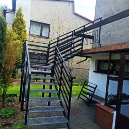 Zewnętrzne schody metalowe z kratowymi stopniami i czarną balustradą prowadzące na balkon, widok z dołu na tle domu z ogrodem.