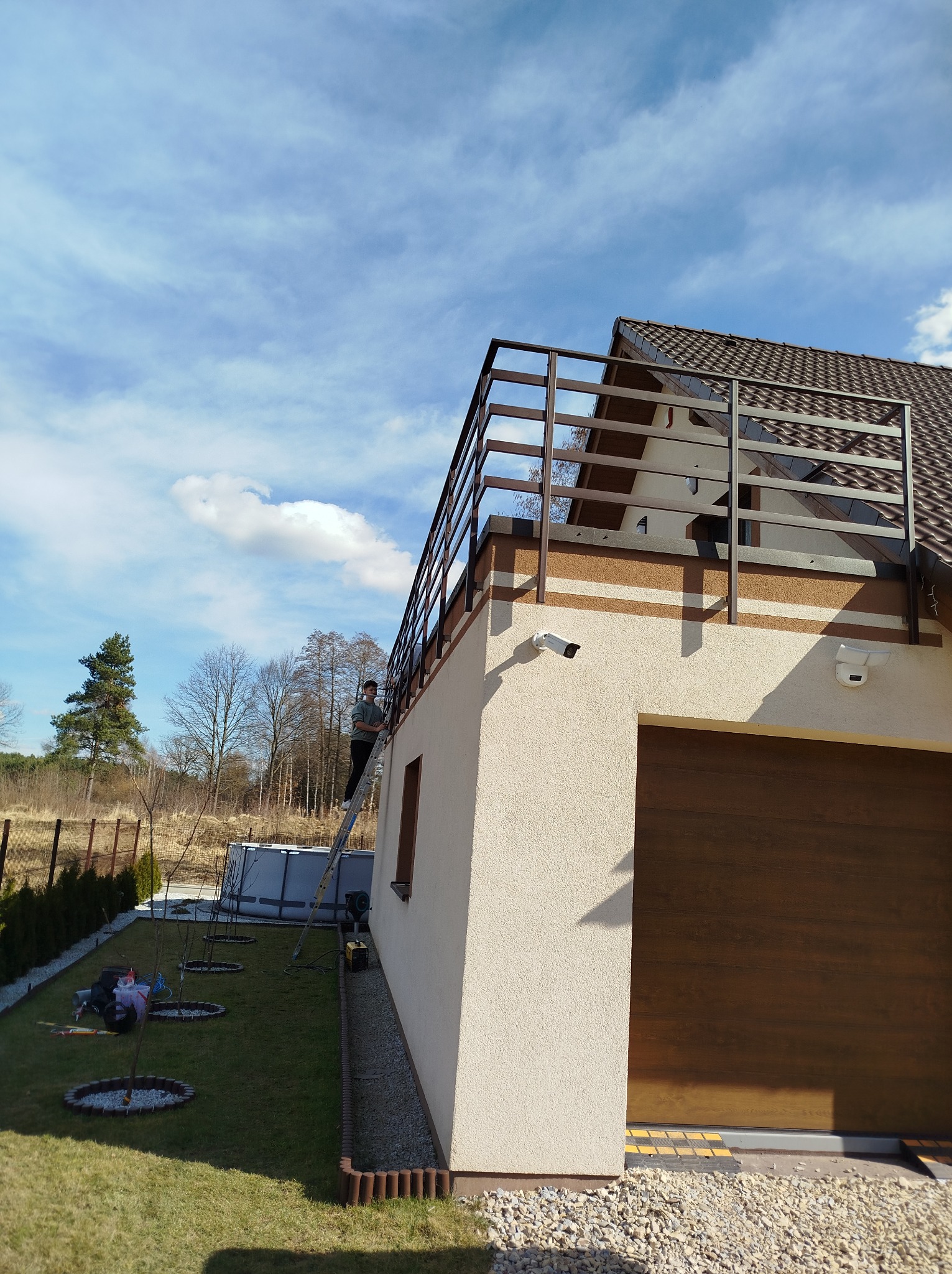Mężczyzna na drabinie przy brązowej, metalowej balustradzie balkonowej nowoczesnego domu z garażem; widoczne kamery monitoringu i świeżo posadzone drzewka.