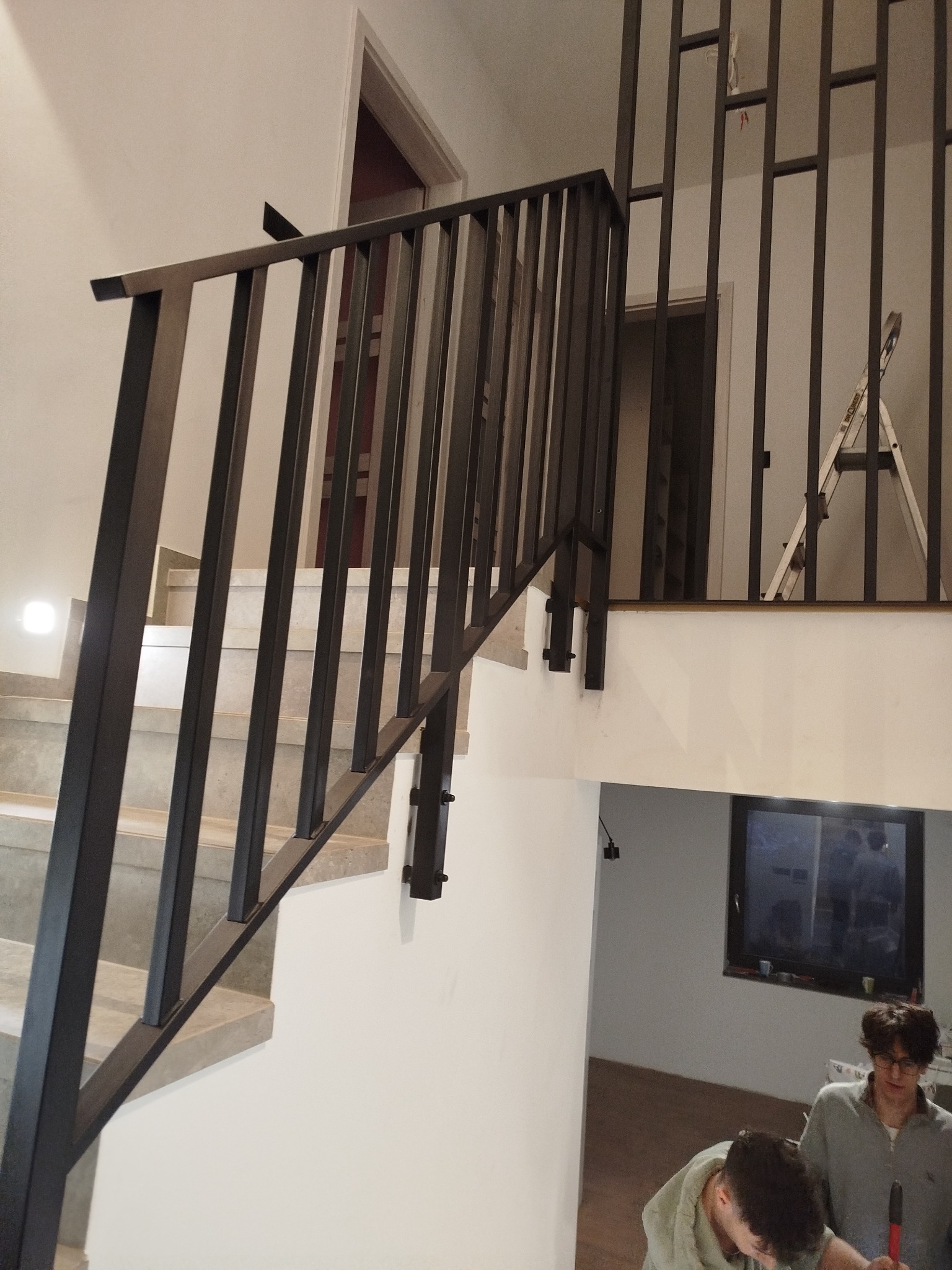 Nowoczesna, czarna balustrada schodowa z pionowymi szczeblami, widok na schody z jasnego kamienia i fragment pomieszczenia z oknem oraz dwoma osobami w tle.