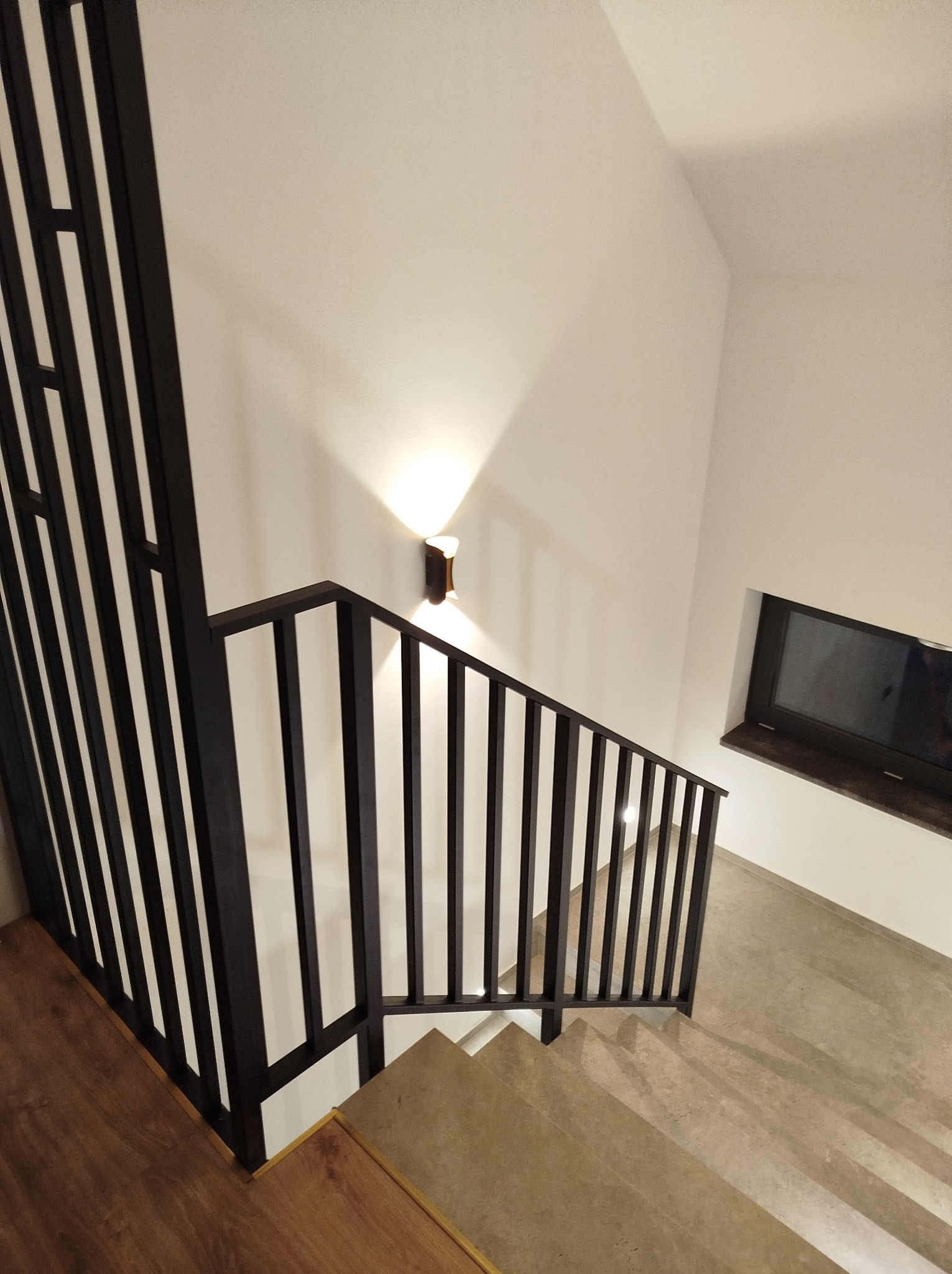 Nowoczesna klatka schodowa z ciemną, metalową balustradą o minimalistycznym designie, oświetlona kinkietem, widok z góry na schody z betonową wylewką.